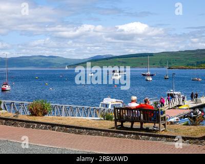 Ein Paar, das an einem schönen Tag auf einer Bank am Meer sitzt und den Blick über die Tobermory Bay genießt, Isle of Mull, Inner Hebrides, Schottland, Großbritannien. Stockfoto