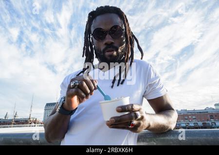 Ein junger afrikanischer Mann mit Bart- und Dreadlocks-Porträt, weiß gekleidet mit dunkler Brille, steht draußen und genießt eine kleine Tasse IK Stockfoto