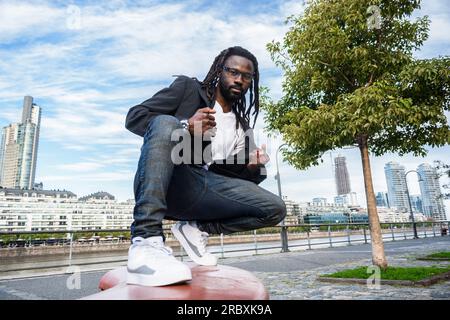Modischer und stilvoller junger schwarzer Mann afrikanischer ethnischer Herkunft mit Dreadlocks, Bart und Sonnenbrille, er trägt lässige Kleidung und posiert Crouchi Stockfoto
