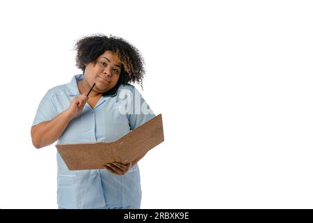 Afro-Ärztin in blauer Uniform, die auf die Kamera schaut, am Telefon spricht und denkt, den Stift auf ihr Kinn zu legen, während sie Notizen macht, weiß Stockfoto