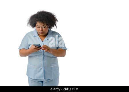 Junge Afro-Latina-Medizinerin venezolanischer ethnischer Herkunft in blauer Uniform, steht und überprüft die Nachrichten auf ihrem Handy, weißer Hintergrund mit Kopie Spa Stockfoto