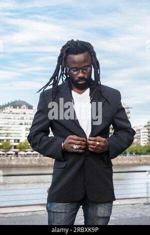 Vertikales Porträt eines modischen jungen afrikanischen Geschäftsmanns mit Dreadlocks, Bart und dunkler Brille, der draußen steht und einen Blazer zuknöpft. Stockfoto