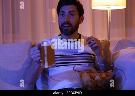 Mann isst Chips und trinkt Bier, während er nachts auf dem Sofa fernsieht. Schlechte Angewohnheit Stockfoto