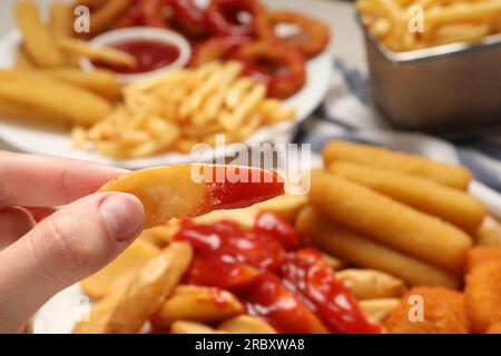 Eine Frau, die leckeren gebackenen Kartoffelkeil mit Ketchup in der Nähe verschiedener Snacks am Tisch hält, Nahaufnahme Stockfoto