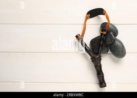 Schwarze Steinschleuder mit Steinen auf weißem Holztisch, flach liegend. Platz für Text Stockfoto