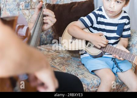 Kaukasisches Kind spielt und macht Musikakkorde mit kleiner Gitarre oder Ukulele, Nahaufnahme. Erwachsene unterrichten das Kind entspannt. Stockfoto