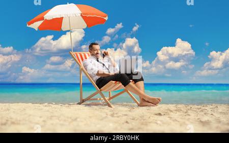 Bussinesman sitzt auf einem Liegestuhl unter dem Sonnenschirm und benutzt am Strand einen Laptop und ein Smartphone Stockfoto