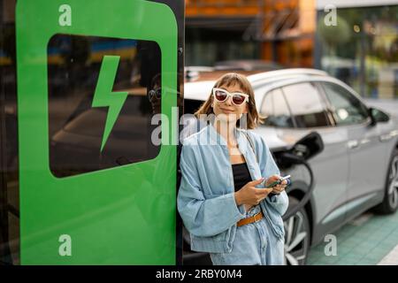 Eine Frau mit einem Smartphone, die darauf wartet, dass ihr Elektroauto aufgeladen wird Stockfoto
