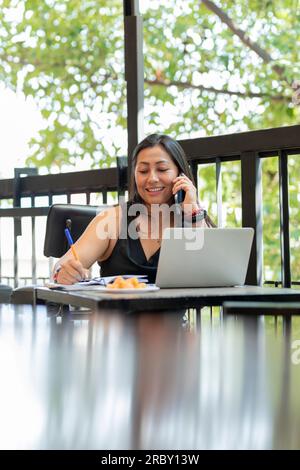 Eine Mexikanerin macht sich Notizen in ihrem Tagebuch, während sie in einem Café im Freien telefoniert. Eine Frau, die in einem Kaffee arbeitet Stockfoto