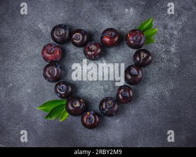 Süße Pflaumen auf dunklem Hintergrund. Frische blaue Pflaumen mit Blättern. Kulinarische Frucht im Hintergrund. Sommerpostkarte. Die Pflaumen werden im Garten geerntet und liegen in Herzform, Draufsicht Stockfoto