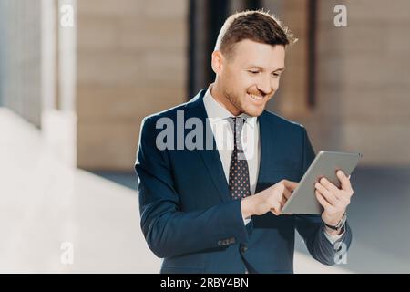 Erfolgreicher männlicher Unternehmer im formellen Anzug, hält modernes Tablet, prüft die Dokumentation online, liest die notwendigen Informationen für die Erstellung von Geschäftsberichten, Stockfoto