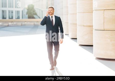 Wohlhabender männlicher Geschäftsinhaber in formellen Anzügen, führt Telefongespräche mit Geschäftspartnern, während er zu einem Meeting in urbaner Umgebung geht, zufrieden Stockfoto