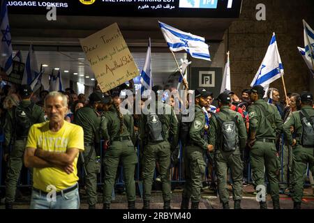 Israelische Grenzpolizisten stellen sich vor Demonstranten auf, die während einer Demonstration gegen die Justizreform am Flughafen Ben Gurion mit israelischer Flagge wedeln. Stockfoto