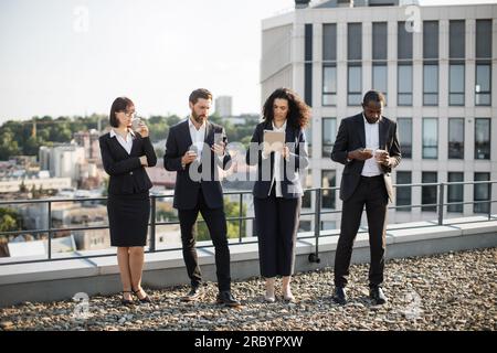 Erfolgreiche männliche und weibliche Kollegen, die während der Kaffeepause auf freiem Raum in sozialen Medien scrollen. Multikulturelles Spezialistenteam mit kurzen Pausen nach langen Verhandlungen. Stockfoto