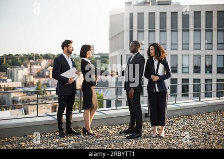 Birassische Geschäftspartner geben sich die Hand, um die Vereinbarung gegenseitiger Investitionen in Neubauten zu bestätigen. Vier Unternehmer in Anzügen treffen sich auf der Büroterrasse. Konzept von Teamarbeit und Zusammenarbeit. Stockfoto