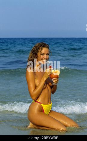 Fröhliche, hübsche junge Frau, die auf die Kamera schaut und Fruchtcoktail trinkt; Meerwasser und blauer Himmel auf dem Hintergrund gesund attraktive Silhouette Körper lächelnd Stockfoto