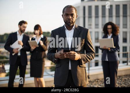 Porträt eines erfolgreichen afroamerikanischen Unternehmers, der sein Handy in den Händen hält und vor der Kamera lächelt. Geschäftspartner mit gemischter Staatsangehörigkeit, die verwenden Stockfoto