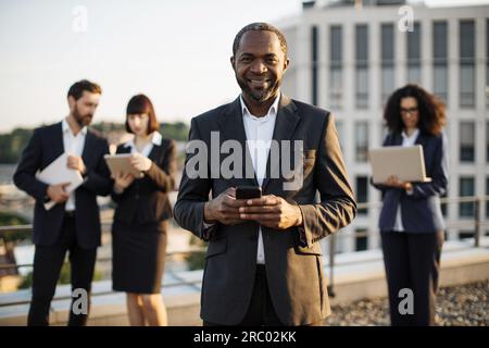 Porträt eines erfolgreichen afroamerikanischen Unternehmers, der sein Handy in den Händen hält und vor der Kamera lächelt. Geschäftspartner mit gemischter Staatsangehörigkeit, die verwenden Stockfoto