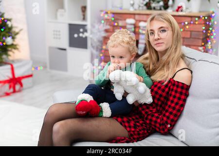 Eine junge blonde Frau mit Brille sitzt auf einer grauen Couch und hält ein Baby auf dem Schoß Stockfoto