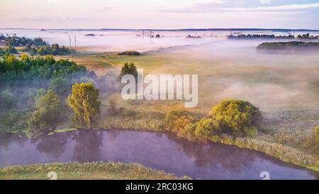 Atemberaubender Blick auf den Sonnenaufgang im Sommer. Ländliche Landschaft, der Fluss schlängelt sich durch üppige grüne Bäume. Morgen-Foggy-Szene. Ruhige und friedliche Atmosphäre. Kleiner Bauernhof Stockfoto