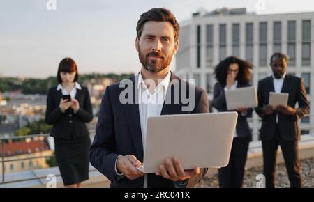 Porträt eines fröhlichen weißlichen Geschäftsmanns, der mit einem Laptop in der Hand in die Kamera schaut und lächelt. Eine vielfältige Gruppe von Spezialisten, die mit Mod in Anzügen gekleidet sind Stockfoto
