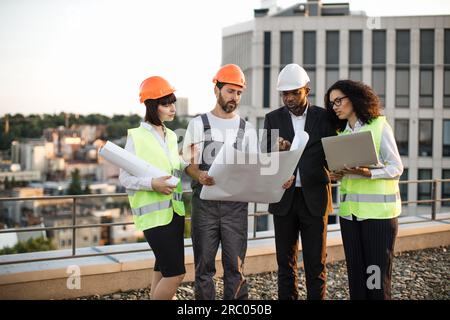 Gruppe von vier multiethnischen Personen in Anzügen und Helmen, die Brainstorming für ein gemeinsames Projekt eines neuen Immobiliengebäudes durchführen. Bauteam, das mit blu zusammenarbeitet Stockfoto