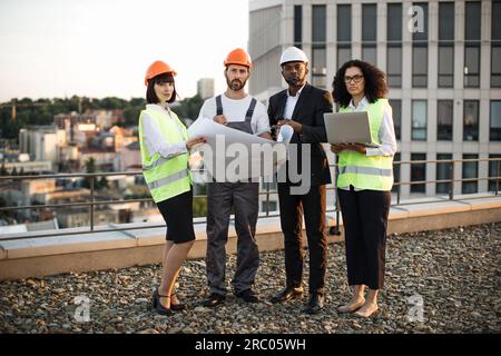 Das ganze Porträt eines multiethnischen Arbeitsteams, das auf einer Terrasse steht und einen fantastischen Blick auf die Stadt hat und in die Kamera schaut. Vier internationale Stadtplaner in Schutzkleidung mit Layouts und Laptop. Stockfoto