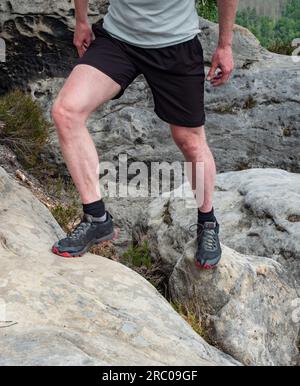 Details zu Wanderbeinen in schwarz-roten Wanderschuhen auf dem Berggipfel. Füße in Wanderschuhen und Beine leichte Hosen auf dem Hintergrund der Gipfel Stockfoto