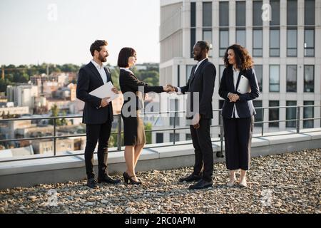 Birassische Geschäftspartner geben sich die Hand, um die Vereinbarung gegenseitiger Investitionen in Neubauten zu bestätigen. Vier Unternehmer in Anzügen treffen sich auf der Büroterrasse. Konzept von Teamarbeit und Zusammenarbeit. Stockfoto