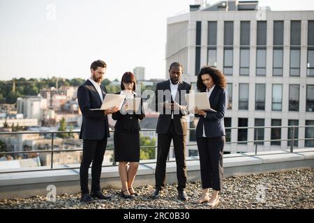 Vier Investoren mit kaukasischer und afroamerikanischer Nationalität in Business Outfit, die auf dem Dach mit technischen Geräten stehen und Probleme bei der Arbeit lösen. Jeder Partner schaut sich sein eigenes Gerät an und schreibt E-Mails. Stockfoto