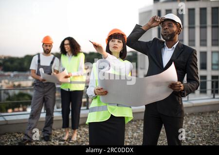 Eine Gruppe unterschiedlicher Ingenieure mit Bauplänen, die auf einem flachen Dach stehen und über den Bauprozess sprechen. Zwei Kollegen mit kaukasischer und afroamerikanischer Nationalität zeigen auf Baustelle. Stockfoto