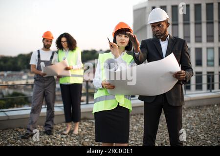 Eine Gruppe unterschiedlicher Ingenieure mit Bauplänen, die auf einem flachen Dach stehen und über den Bauprozess sprechen. Zwei Kollegen mit kaukasischer und afroamerikanischer Nationalität zeigen auf Baustelle. Stockfoto