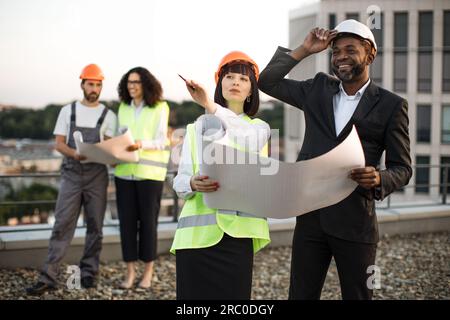 Eine Gruppe unterschiedlicher Ingenieure mit Bauplänen, die auf einem flachen Dach stehen und über den Bauprozess sprechen. Zwei Kollegen mit kaukasischer und afroamerikanischer Nationalität zeigen auf Baustelle. Stockfoto