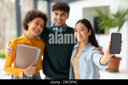 Fröhliche, vielfältige Schüler, Freunde, die sich umarmen, und eine asiatische Dame, die einen leeren Bildschirm auf dem Handy zeigt, eine Werbe-App für Bildung Stockfoto