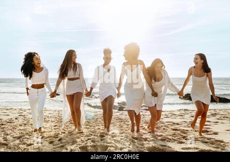 Eine lange Aufnahme von glücklichen Damen, die am Meer spazieren, Händchen halten und lachen, eine Hühnerparty am Strand veranstalten Stockfoto