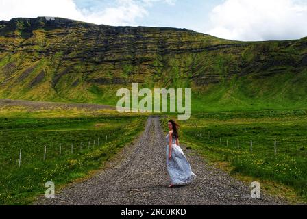 Eine Frau in einem silbernen Kleid steht inmitten der majestätischen grünen Berge nahe Mossfellsbaer, Island. Stockfoto