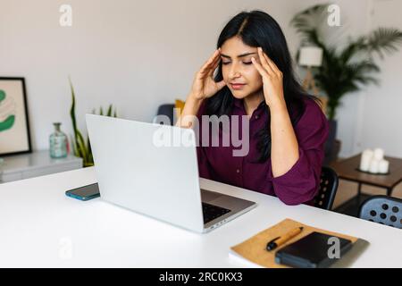 Junge indische Frau, die bei der Arbeit mit einem Notebook Kopfschmerzen oder Migräne hat Stockfoto