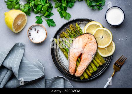 Gedämpfter Lachs mit gebackenem Spargel und Zitrone auf Keramikplatte. Tischhintergrund aus grauem Stein, Draufsicht Stockfoto
