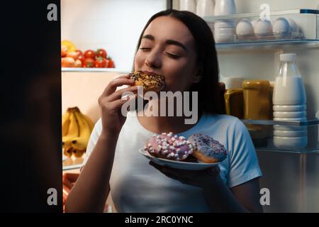 Junge Frau isst Donut in der Nähe des Kühlschranks in der Küche. Schlechte Angewohnheit Stockfoto