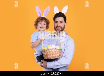 Glücklicher Vater und Sohn mit süßen Hasenohren Stirnbändern auf orangefarbenem Hintergrund. Ein Junge mit einem Osterkorb und bemalten Eiern Stockfoto