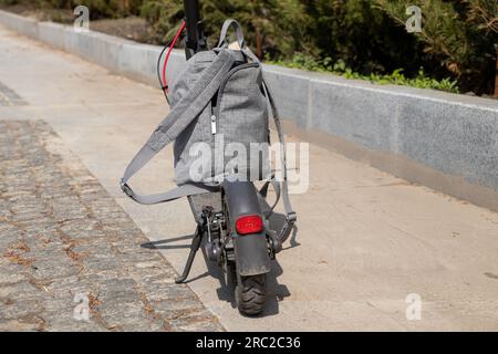 Ein grauer urbaner Rucksack steht auf einem elektrischen Roller im Sommer auf der Straße in der Sonne in der Stadt, im Lifestyle, im Stadtverkehr und in der Mode Stockfoto