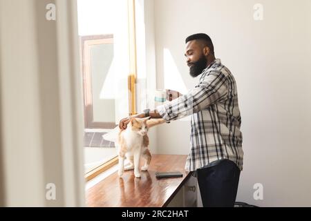 Ein glücklicher afroamerikanischer Mann in einem karierten Hemd, der eine Tasse Kaffee und eine streichelnde Katze hält Stockfoto