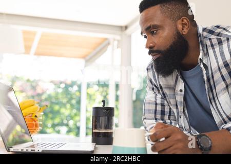 Fokussierter afroamerikanischer Mann mit kariertem Hemd, mit einer Tasse Kaffee und einem Laptop in der Küche Stockfoto