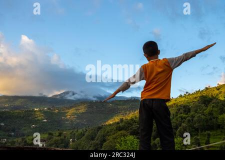 Glücklicher kleiner Junge mit erhobenen Armen, wunderbare Frühlingslandschaft in den Bergen. Grasfeld und sanfte Hügel. Ländliche Landschaft Stockfoto