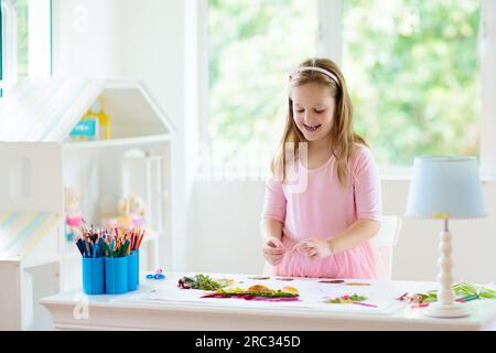 Kind schafft Bild mit bunten Blättern. Kunst und Kunsthandwerk für Kinder. Kleines Mädchen macht Collage Bild mit Regenbogenpflanzen Blatt. Hausaufgaben für die Biologie Stockfoto