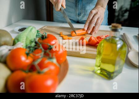 Ernten Sie anonyme Hausfrau, die in der Küche Karotten schneidet, während Sie zu Hause gesunden Salat mit Tomaten und Kräutern zubereiten Stockfoto