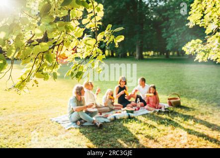 Blütenlindenbaum Ast mit großer Familie, die am Wochenende sonntags auf der Picknickdecke im Stadtpark sitzt. Sie lächeln und lachen und e Stockfoto