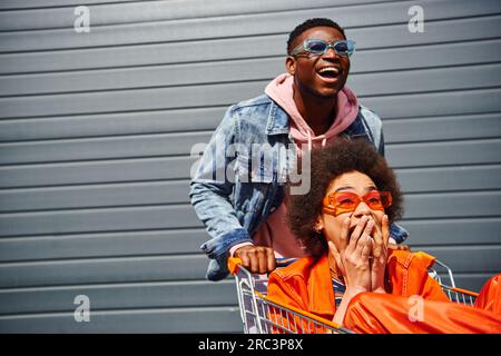 Fröhlicher junger afroamerikanischer Mann mit Sonnenbrille, der Spaß mit seiner verängstigten besten Freundin in trendigen Outfits und einem Einkaufswagen in der Nähe des Gebäudes an der urbanen Straße hat, Stockfoto