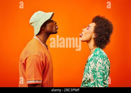 Seitlicher Blick auf junge und trendige afroamerikanische beste Freunde in Sommeroutfits, die ihre Zungen herausragen, während sie isoliert auf orangefarbenen Freunden stehen Stockfoto