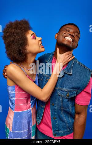 Wütende junge afroamerikanische Frau mit kühnem Make-up berührt den Hals einer fröhlichen besten Freundin in stilvollem Sommeranzug, während sie isoliert auf blau, fa, steht Stockfoto